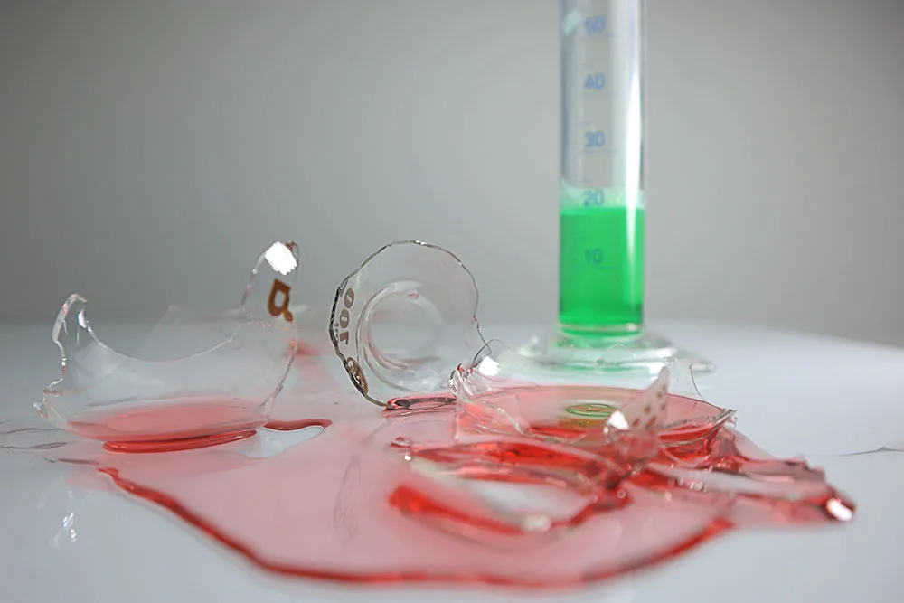 Broken beaker with spilled red liquid, graduated cylinder with green solution, illustrating mk677 mishandling.
